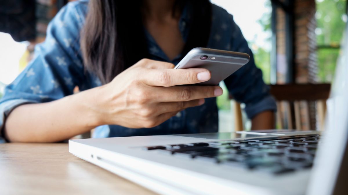 Una mujer consulta un teléfono celular frente a una computadora portátil