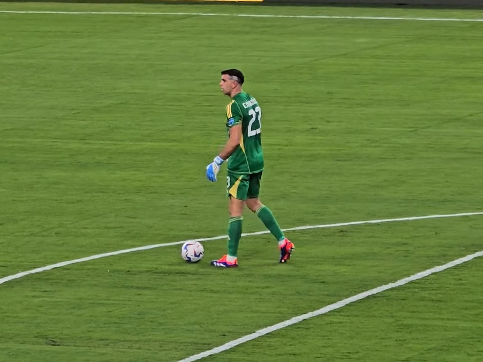 Emiliano Dibu Martínez, con buzo verde número 23 y guantes, camina en el área junto a la pelota durante un partido de la Selección Argentina