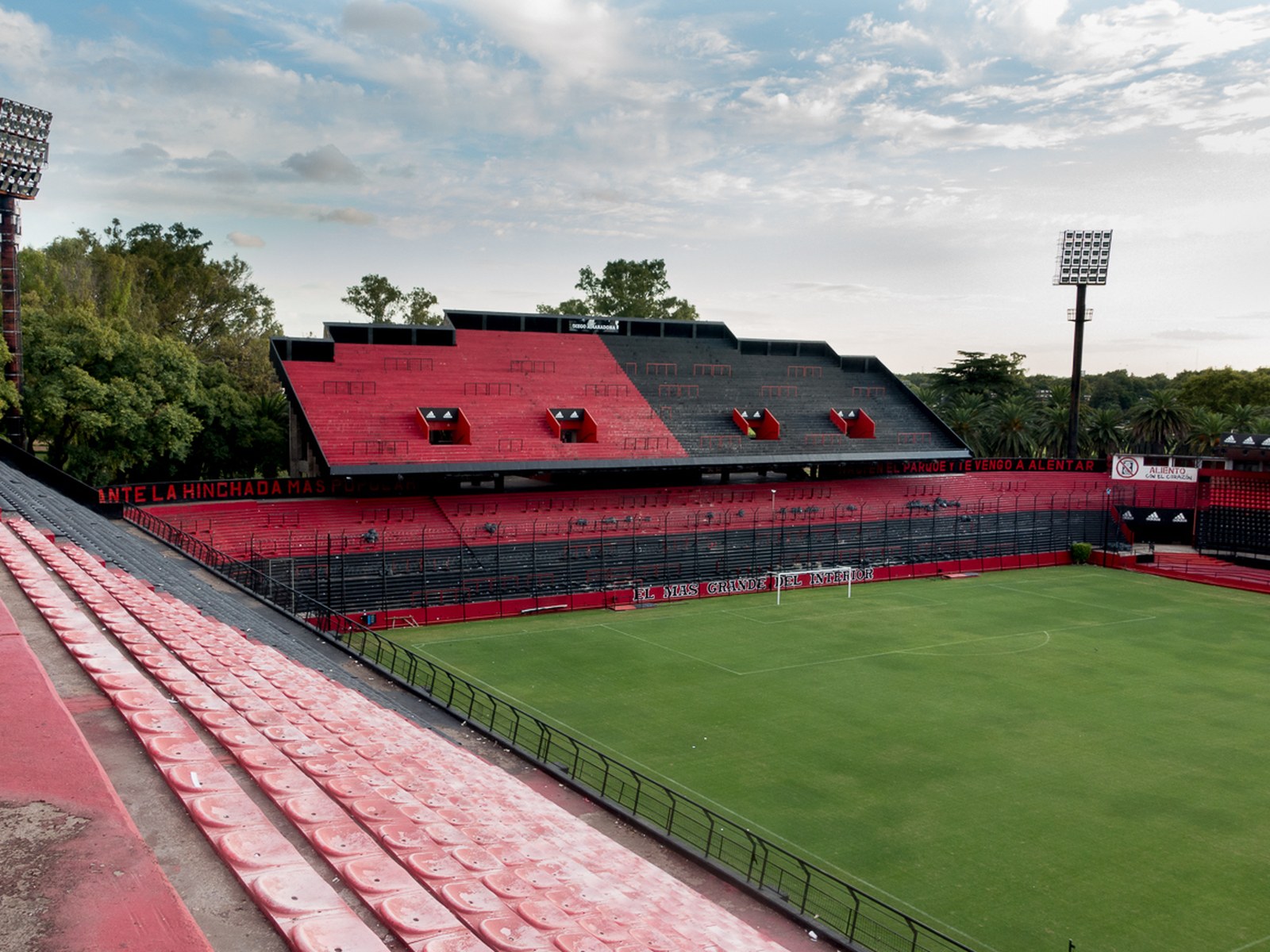 Tribunas rojas y negras del estadio Marcelo Bielsa de Newell's Old Boys, en Rosario, vistas desde lo alto de una platea, con el campo de juego vacío, torres de iluminación y árboles al fondo