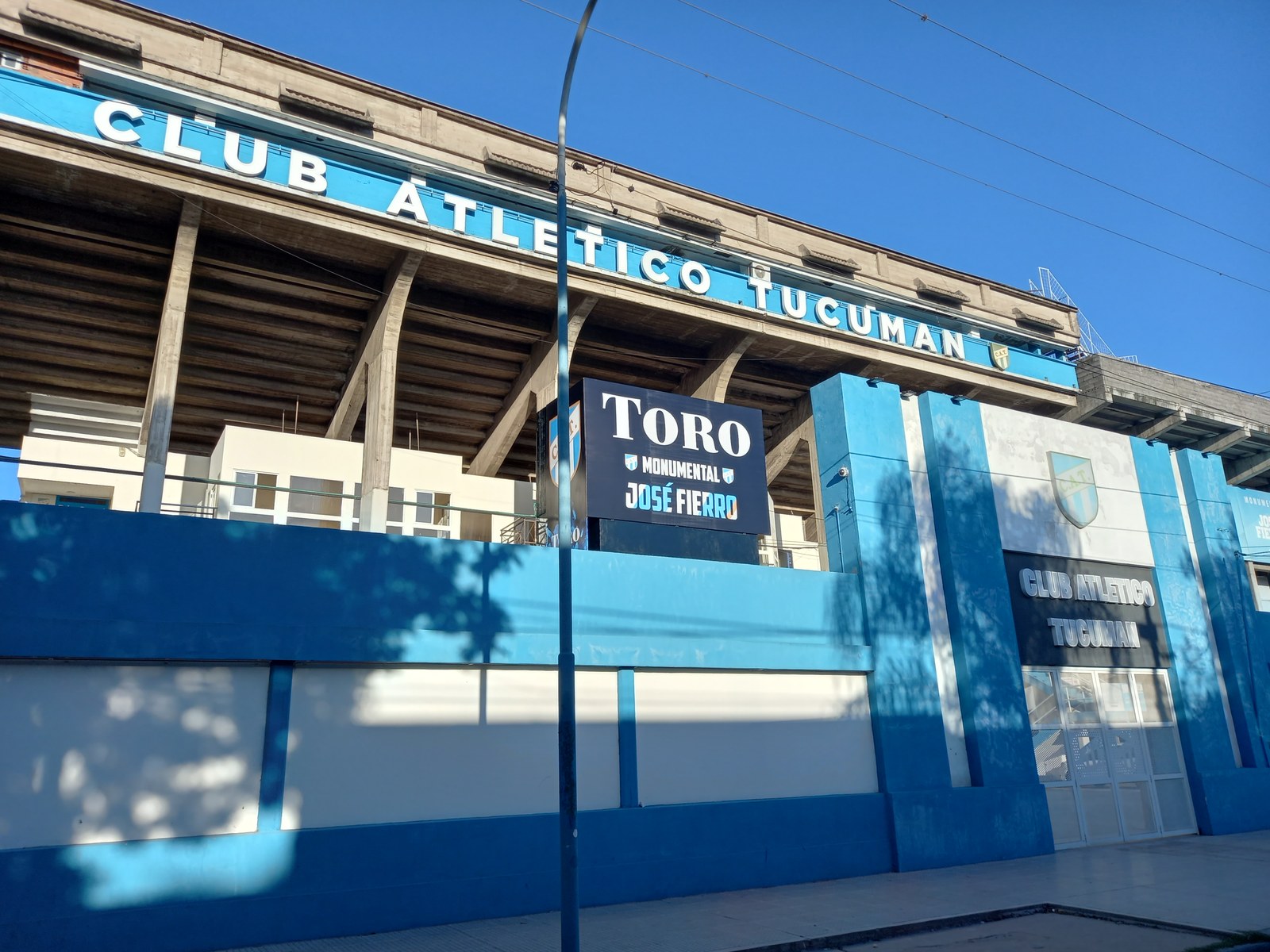 Frente del estadio Monumental José Fierro de Atlético Tucumán, con paredes celestes y blancas, el letrero del club y la tribuna de cemento detrás