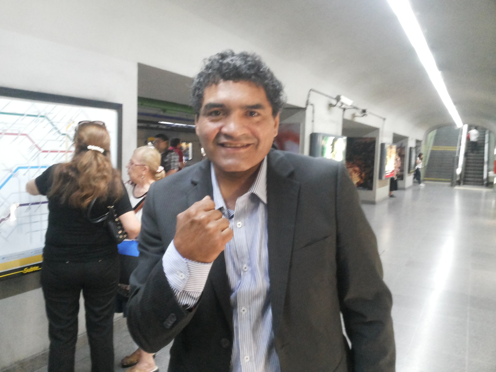 Jorge Locomotora Castro sonriente, de saco oscuro y camisa a rayas, con el puño en alto en una estación de subte