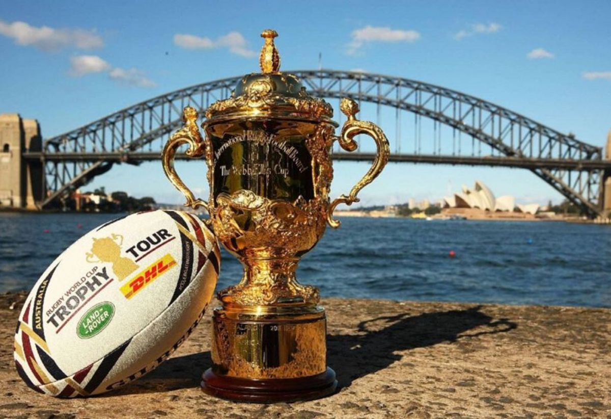La Copa Webb Ellis, trofeo del Mundial de rugby, junto a una pelota sobre la arena, con un puente de fondo