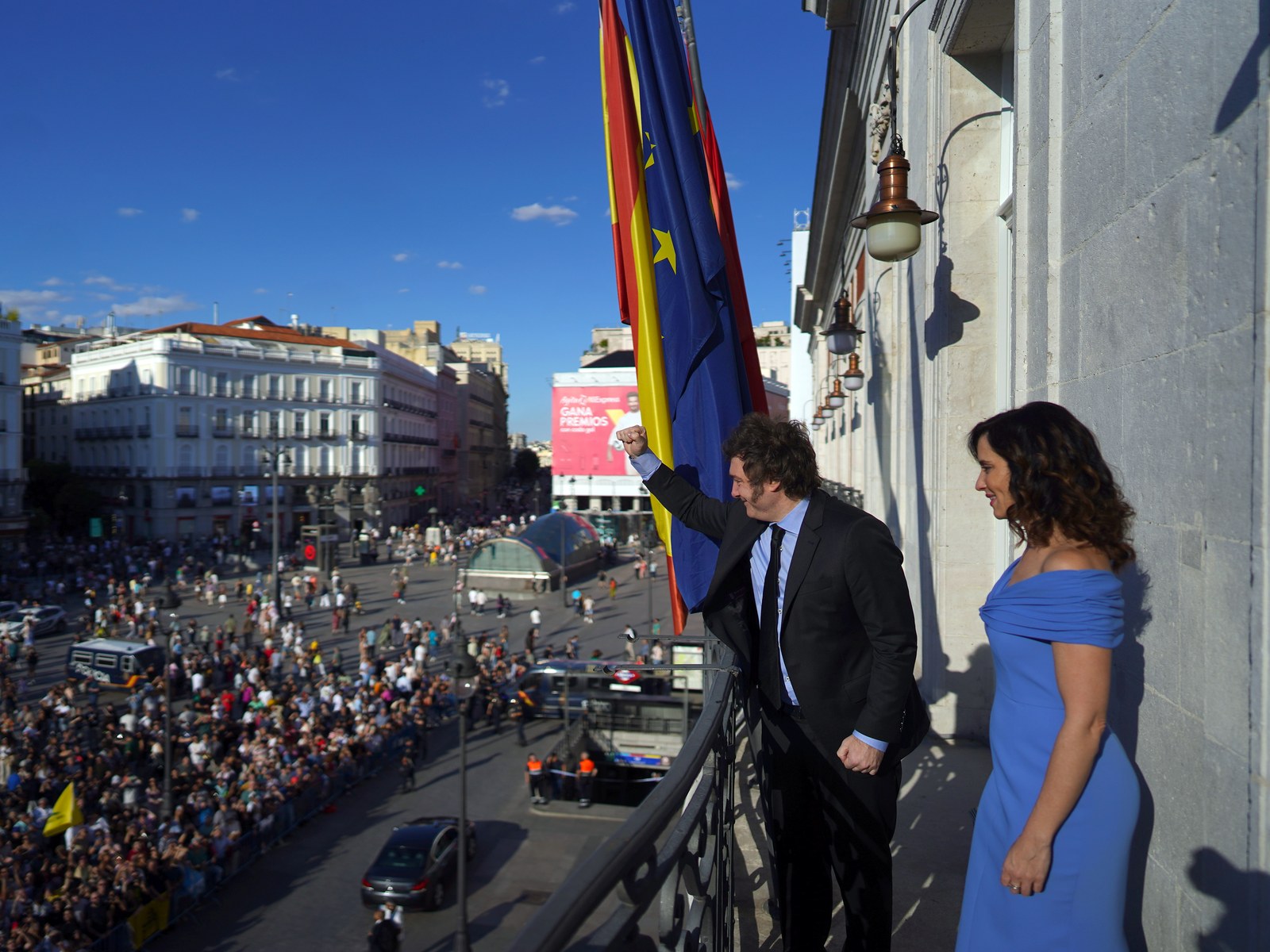 Javier Milei saluda con el brazo en alto desde un balcón junto a Isabel Díaz Ayuso, vestida de azul, con banderas de España y la Unión Europea y una multitud en la plaza de abajo