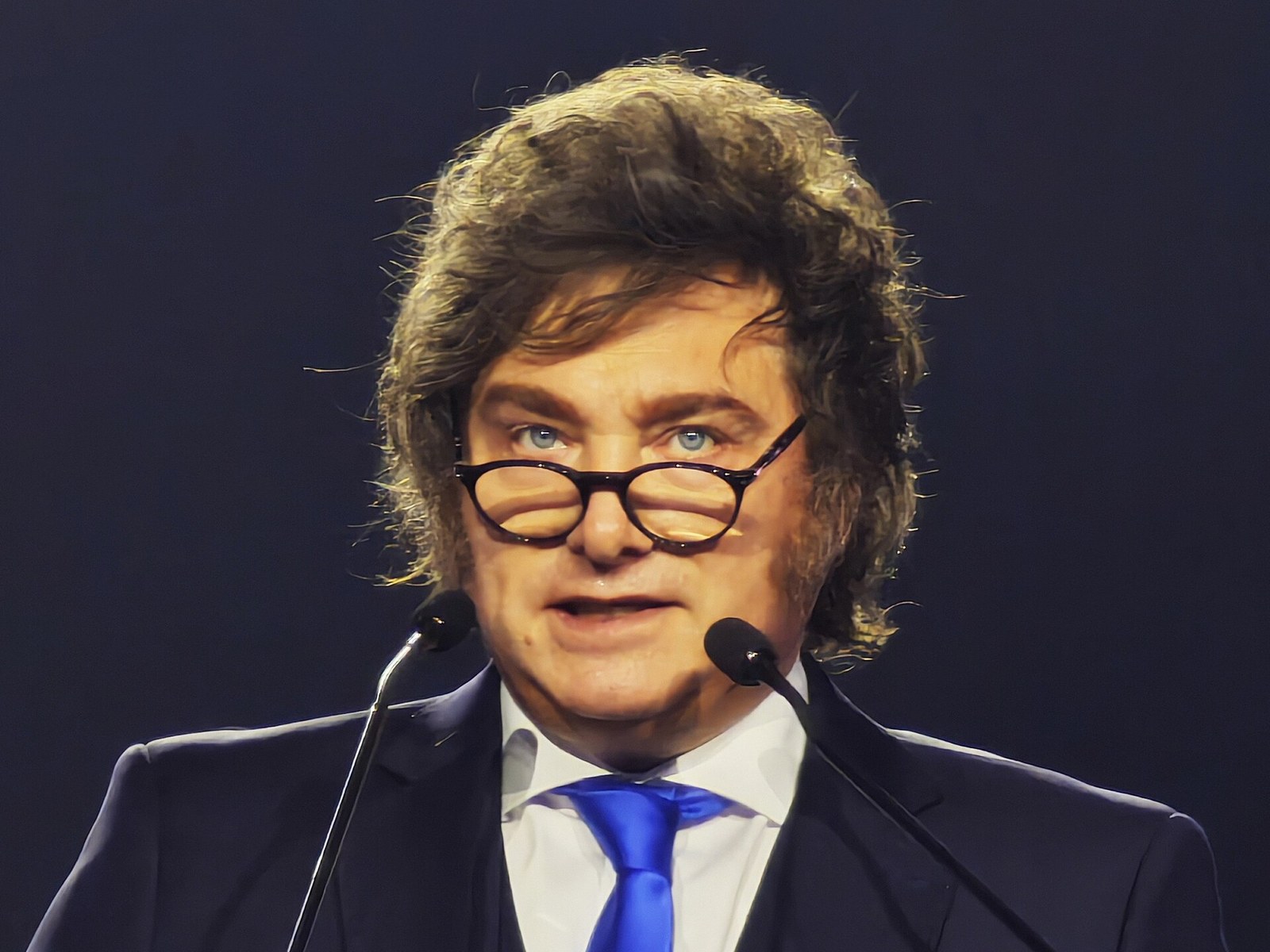 Javier Milei, con anteojos, traje oscuro y corbata azul, habla frente a dos micrófonos sobre un fondo negro