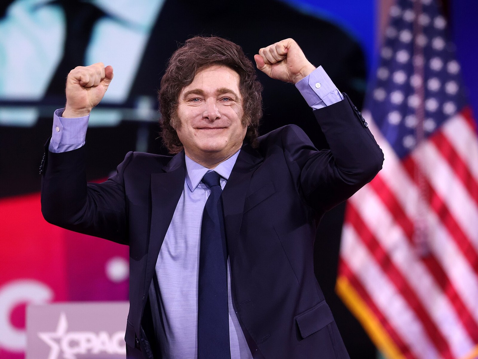 Javier Milei sonriente, con los dos puños en alto en gesto de festejo, de traje oscuro, camisa celeste y corbata azul, con una bandera de Estados Unidos de fondo