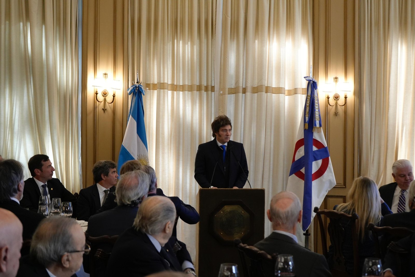 Javier Milei habla desde un atril ante un salón con mesas, junto a una bandera argentina