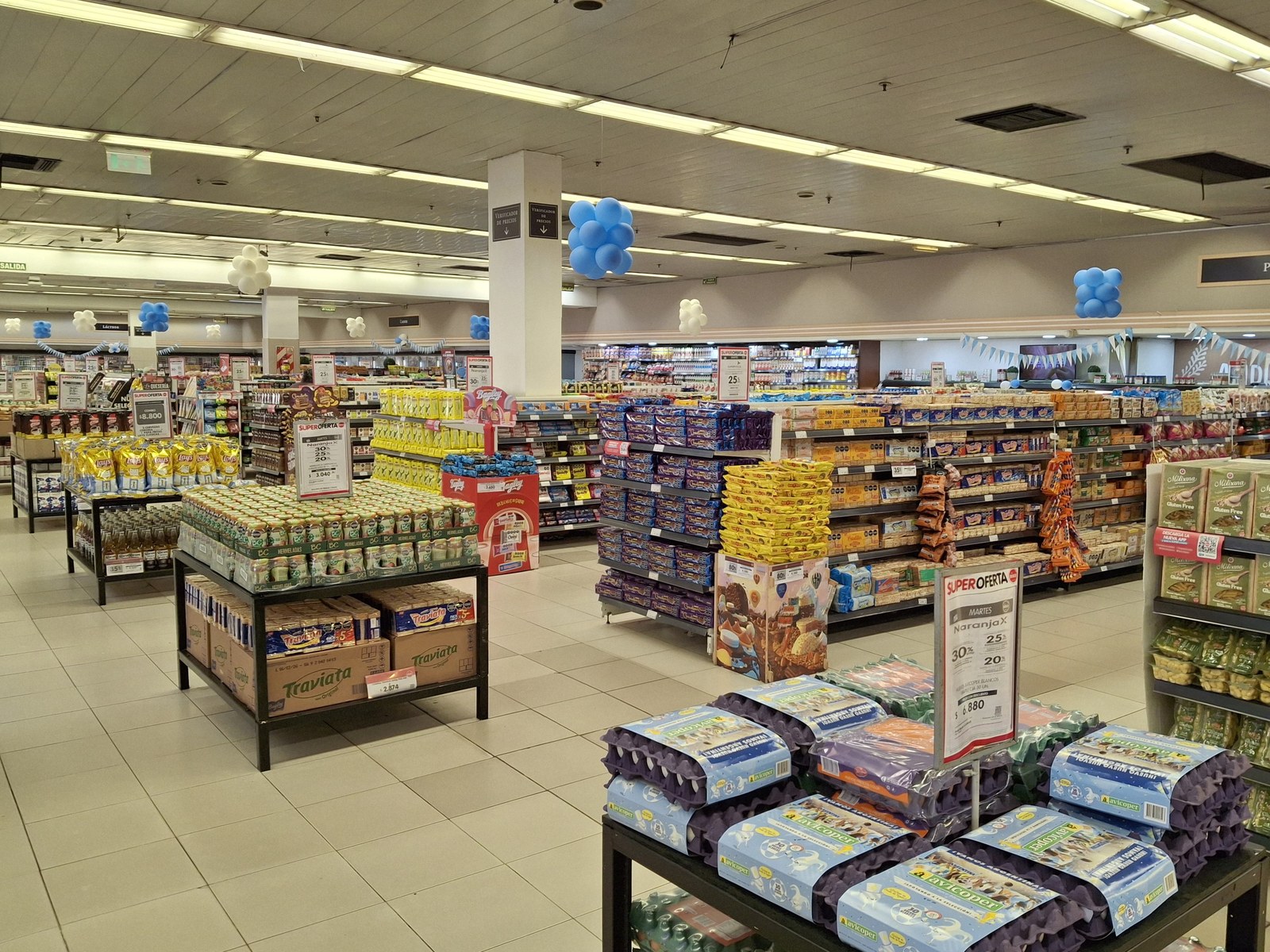 Interior de un supermercado en Morón, provincia de Buenos Aires, con góndolas llenas de productos, carteles de ofertas, maples de huevos en primer plano y globos celestes y blancos colgados del techo