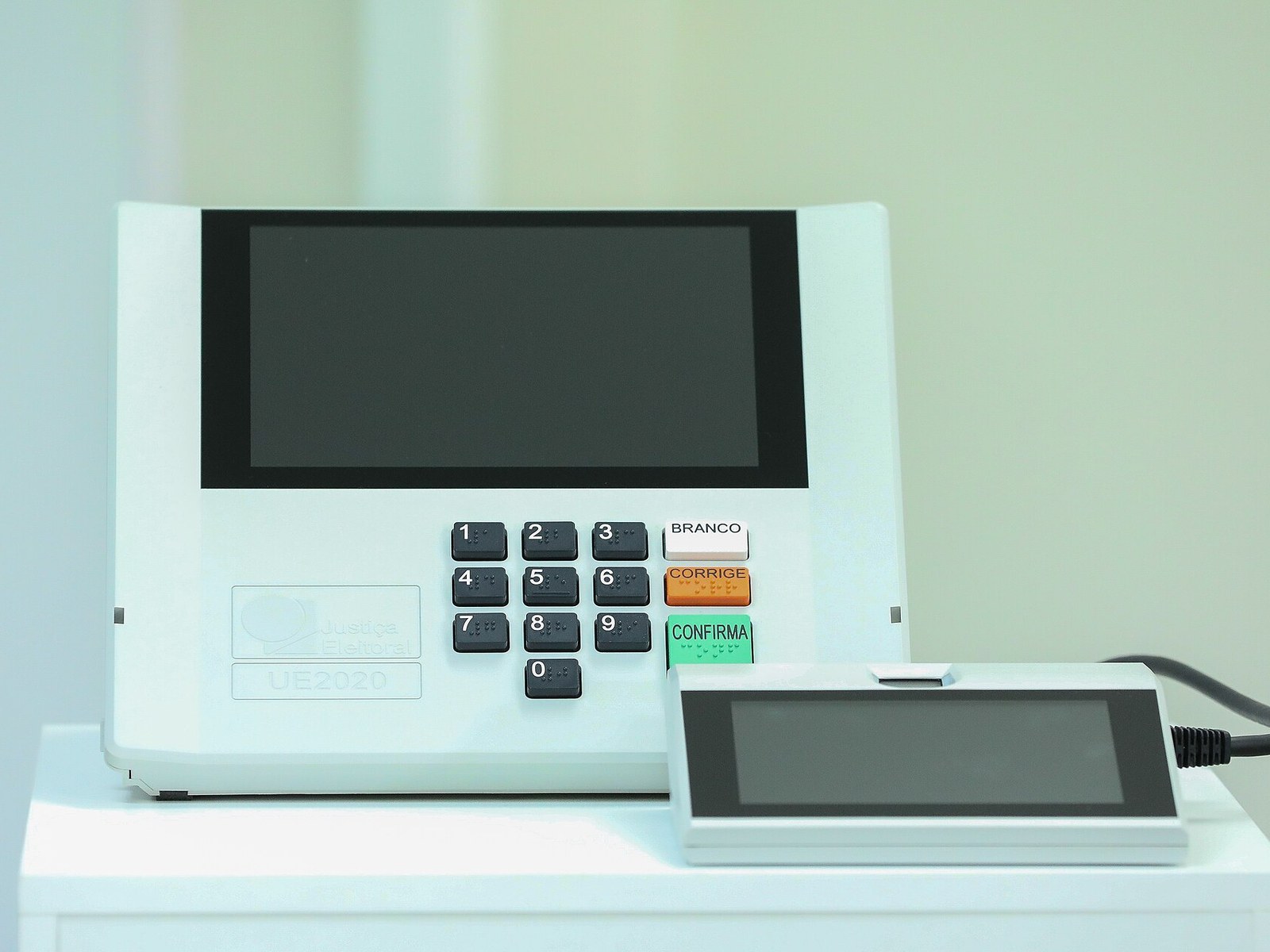 Urna electrónica brasileña modelo UE2020, blanca, con pantalla, teclado numérico y las teclas Branco, Corrige y Confirma, junto al terminal del mesario