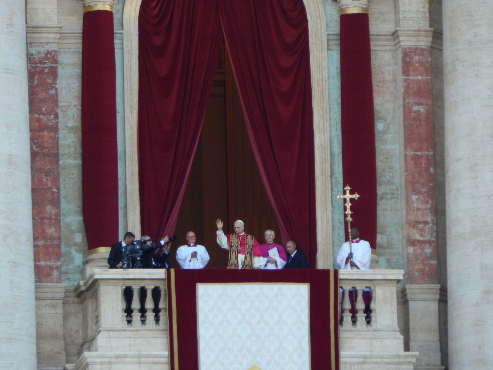 El papa León XIV saluda con la mano en alto desde el balcón central de la Basílica de San Pedro, entre cortinas rojas