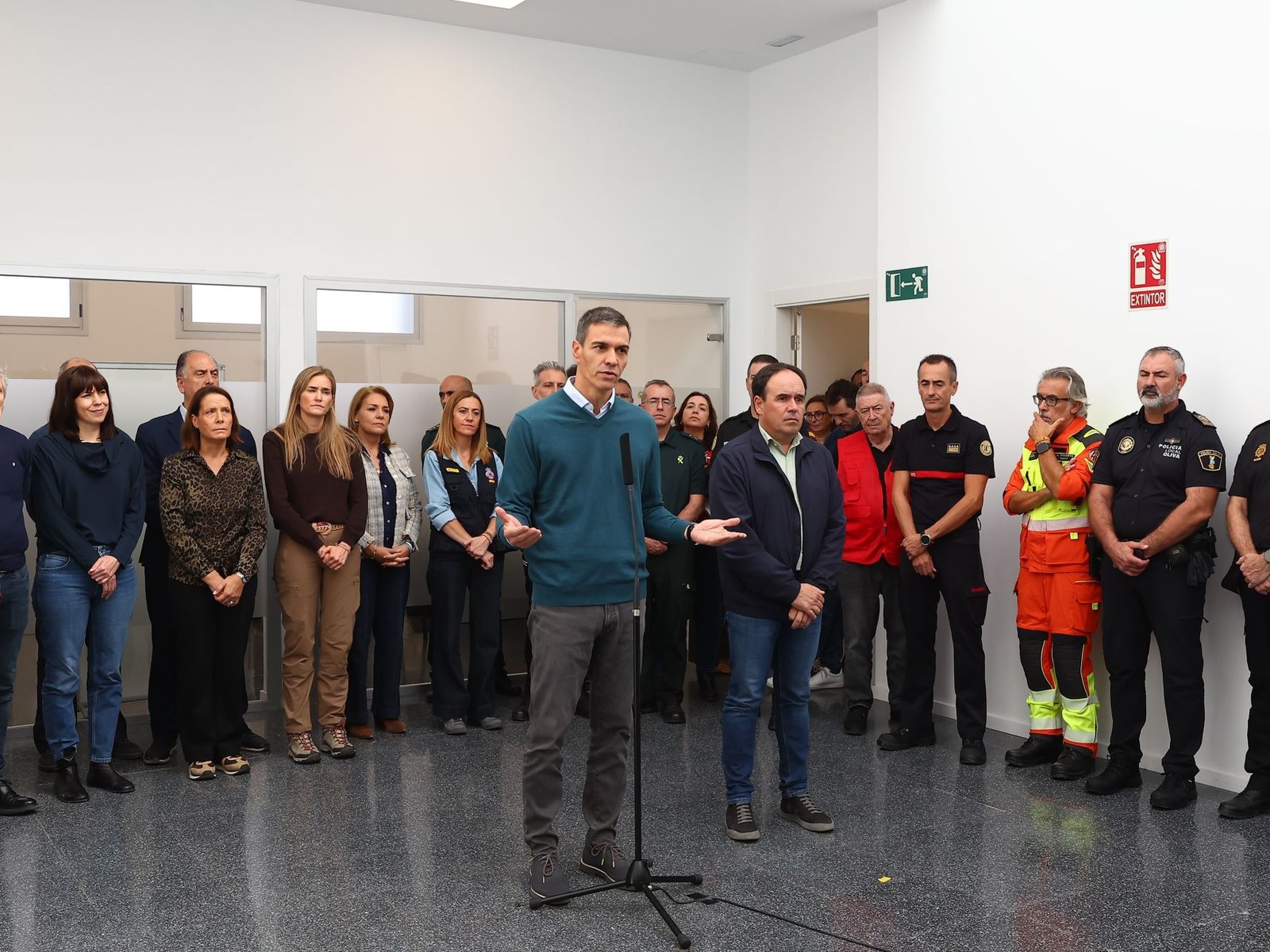 Pedro Sánchez habla frente a un micrófono en una sala blanca, rodeado de autoridades, policías y personal de emergencias con chalecos naranjas, en el puesto de mando de Oliva
