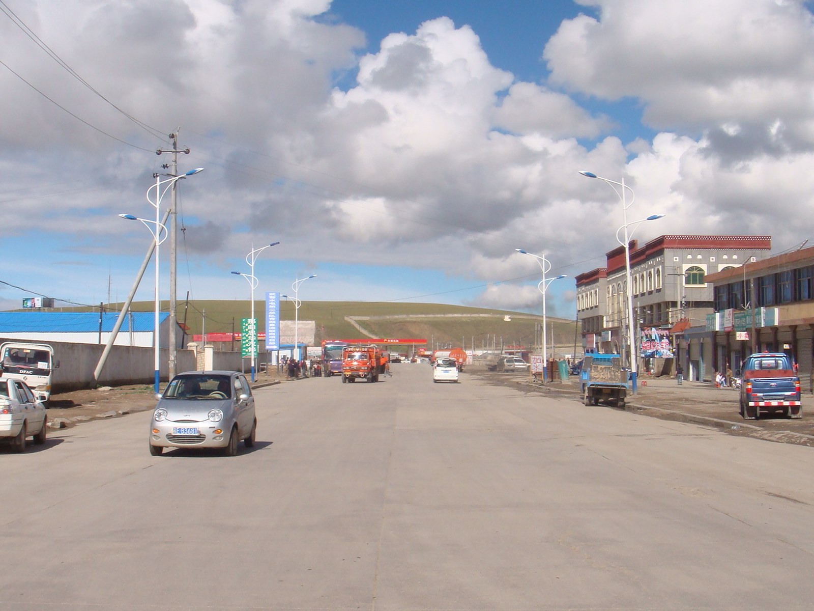 Avenida ancha de la ciudad de Nagqu, en el Tíbet, con autos, edificios bajos y colinas de pastizal al fondo bajo un cielo con nubes