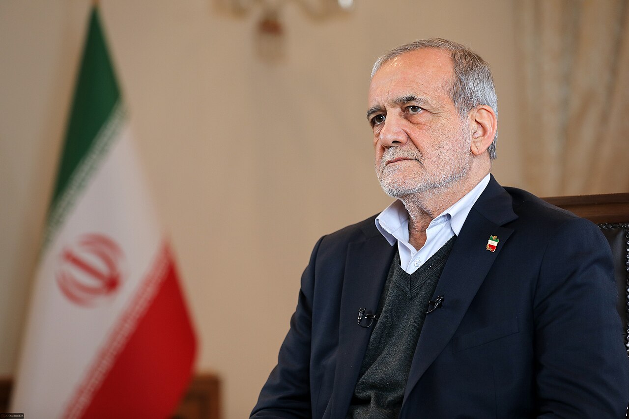 Masud Pezeshkian, de saco oscuro, sentado junto a una bandera de Irán