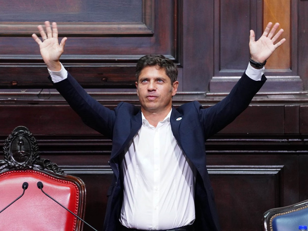 Axel Kicillof, de saco azul oscuro y camisa blanca, con los dos brazos en alto en el recinto de la Cámara de Diputados bonaerense, junto a un sillón de cuero rojo