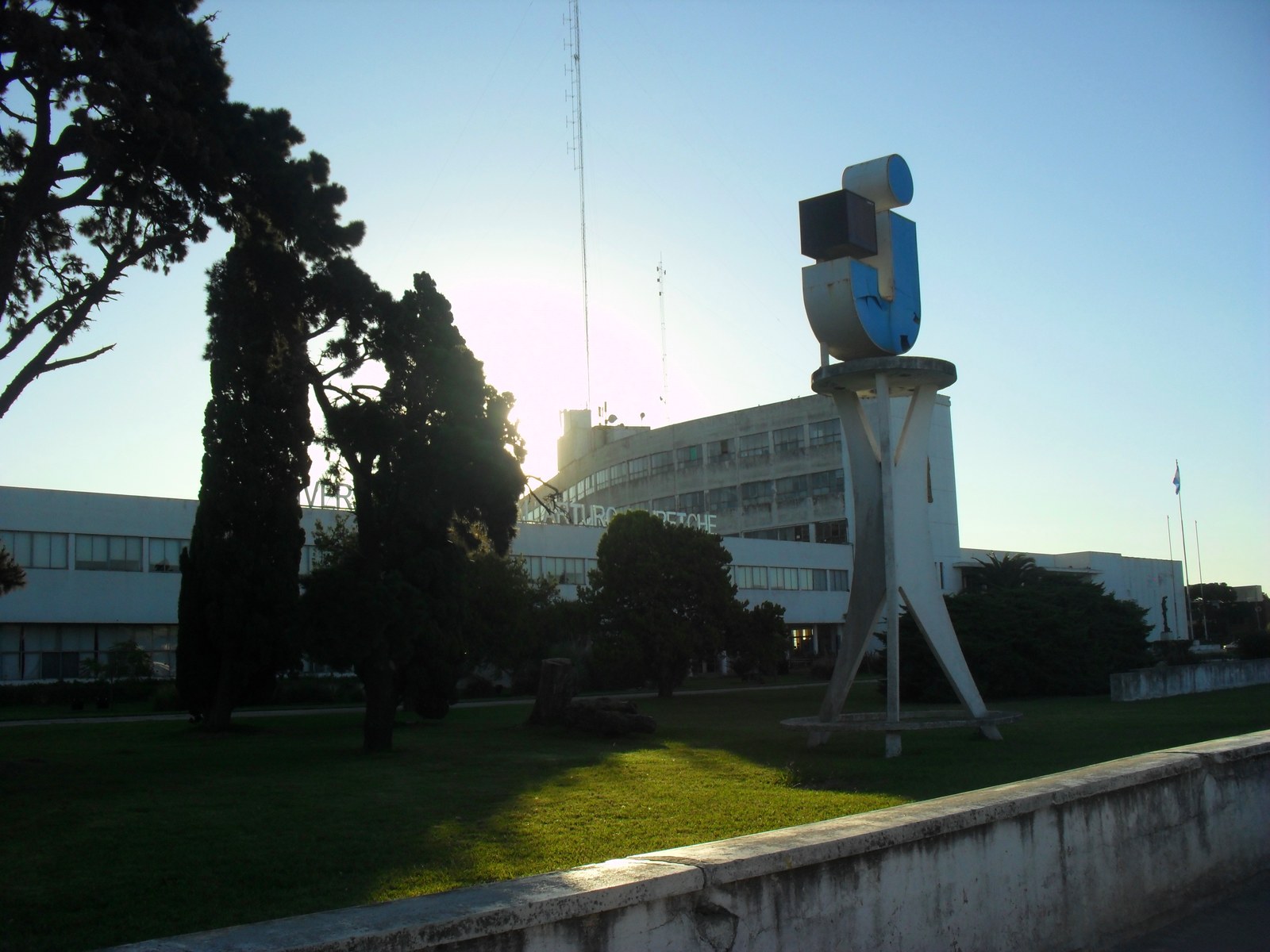 Frente de la Universidad Nacional Arturo Jauretche en Florencio Varela, con el monumento de la letra U azul, árboles y el edificio blanco al fondo a contraluz