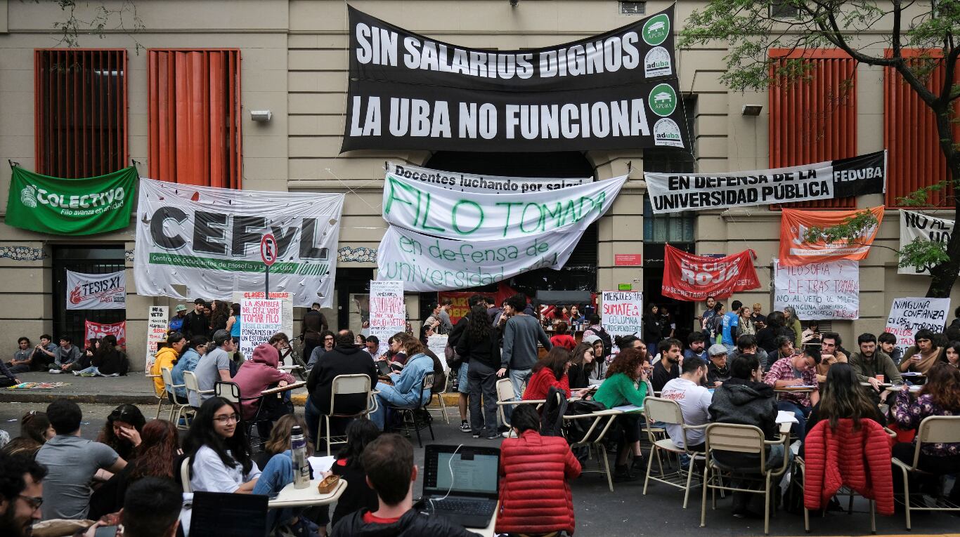 Estudiantes sentados en mesas y sillas en la calle frente a la Facultad de Filosofía y Letras de la UBA, con carteles en defensa de la universidad pública