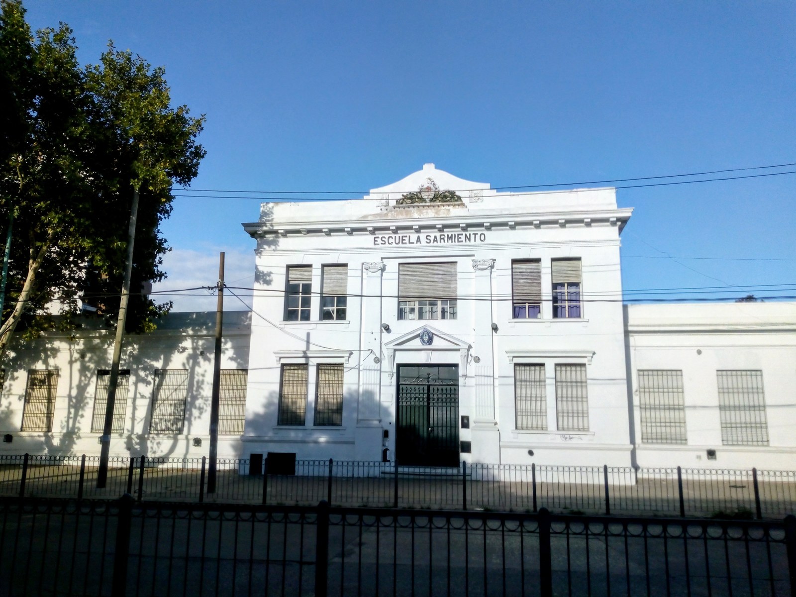 Fachada blanca de la Escuela N° 11 Sarmiento sobre la avenida San Martín, en Florencio Varela, con rejas negras al frente, un árbol y cielo celeste