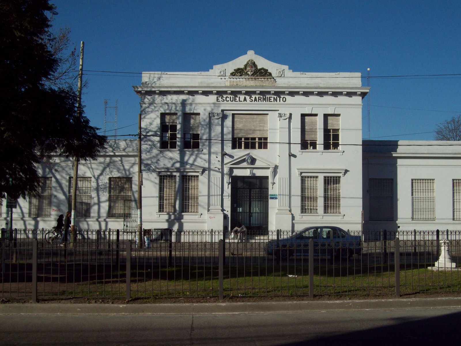 Fachada blanca de la Escuela Sarmiento de Florencio Varela, con su cartel en lo alto, rejas al frente y un cielo despejado