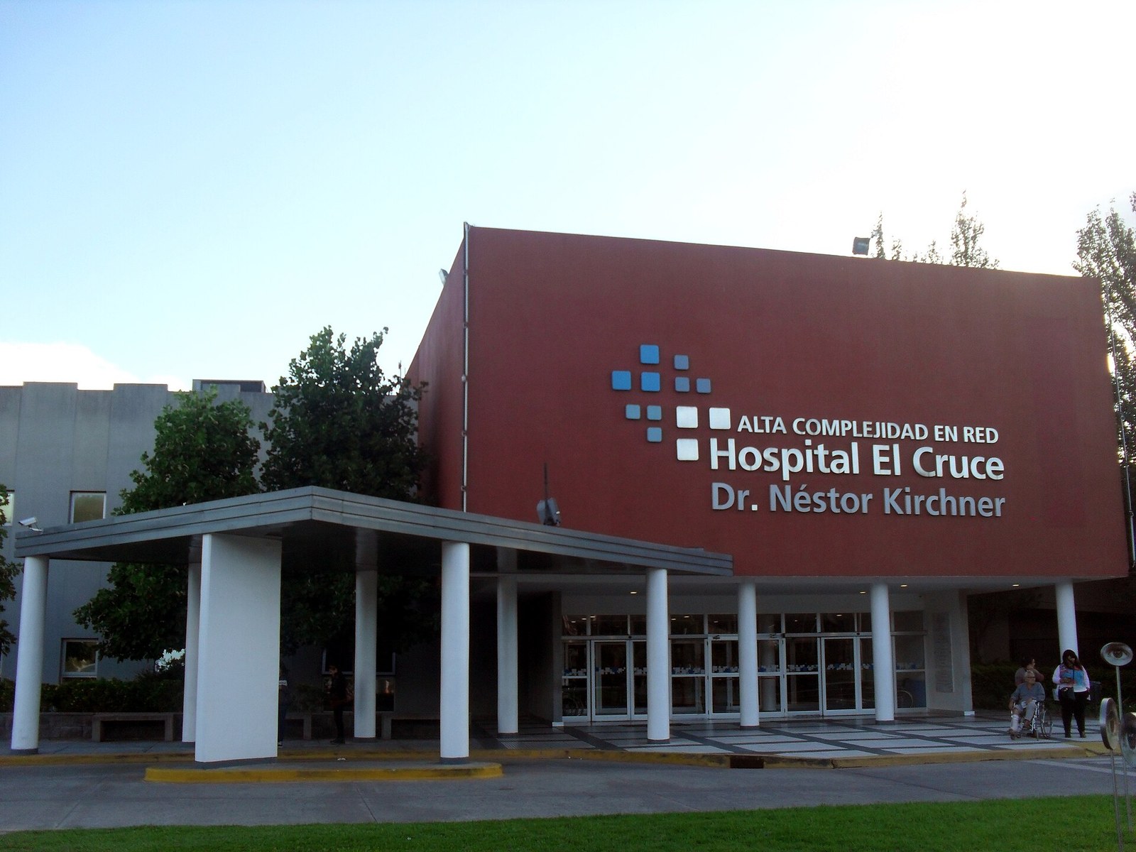 Frente del Hospital El Cruce en Florencio Varela, con la fachada roja y el cartel Alta complejidad en red Hospital El Cruce Dr. Néstor Kirchner, la galería de entrada con columnas blancas y el césped adelante