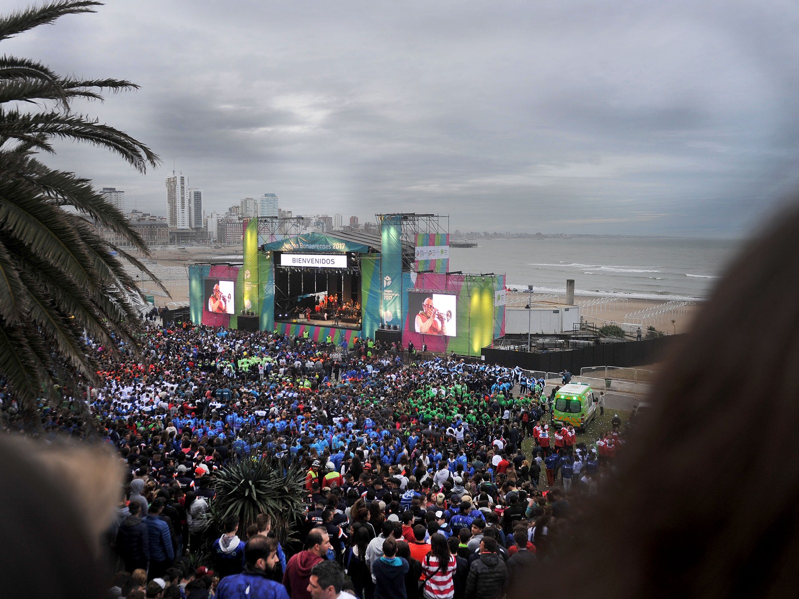 Multitud de chicos y chicas con remeras de colores frente a un gran escenario con pantallas en Las Toscas, Mar del Plata, con edificios y el mar de fondo bajo un cielo nublado