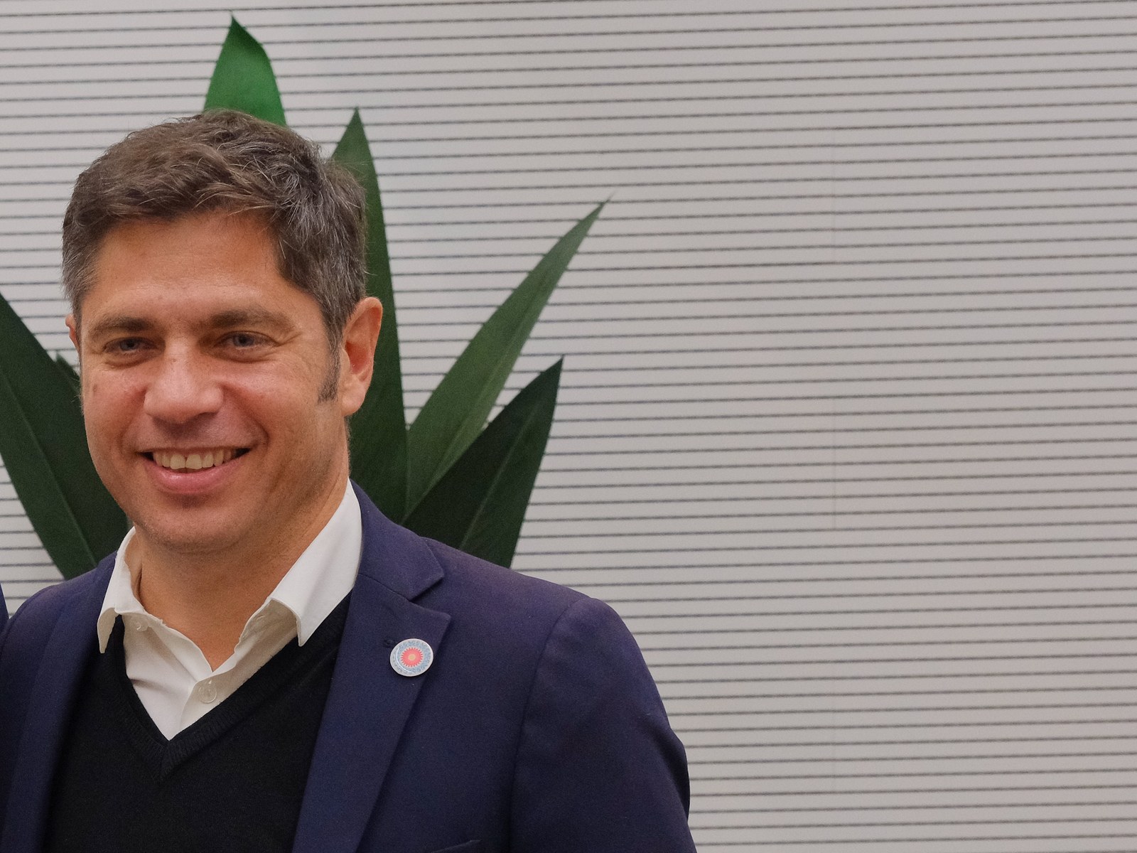 Axel Kicillof sonríe durante una reunión oficial en Barcelona, con saco azul y una planta de fondo