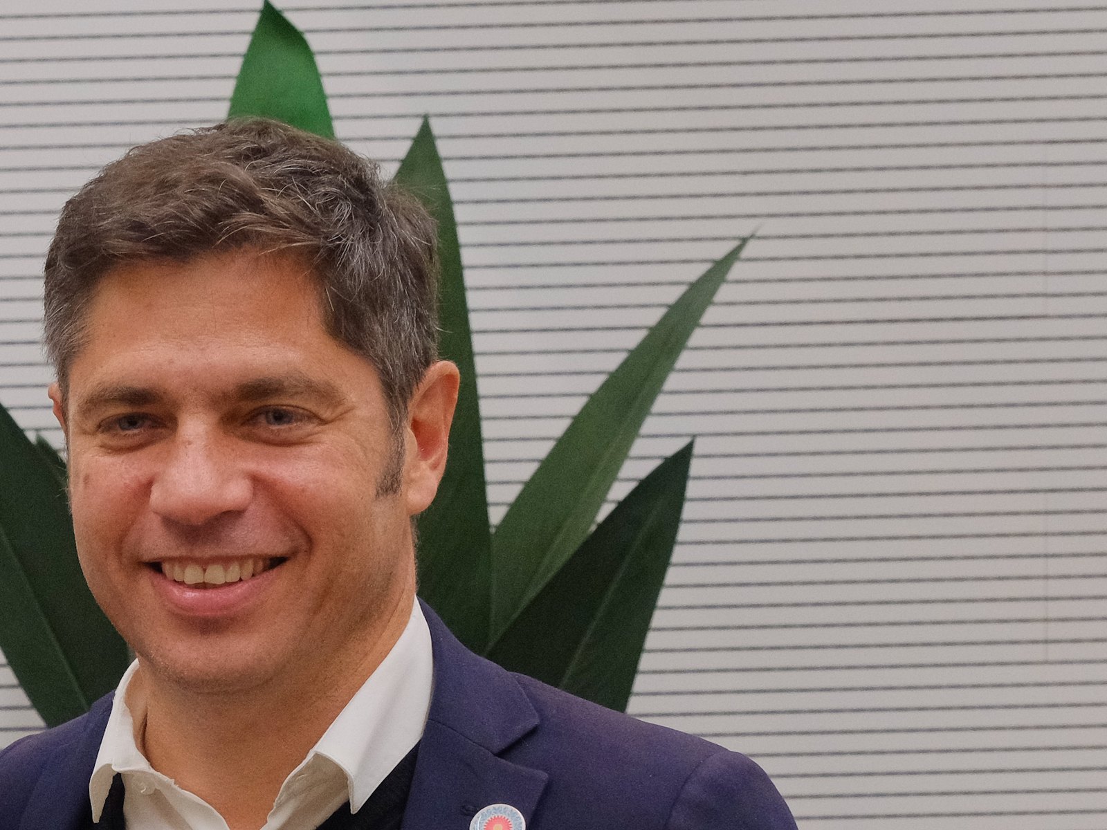 Retrato del gobernador bonaerense Axel Kicillof, sonriente, con saco azul y una planta detrás