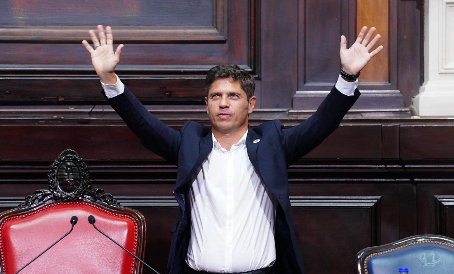 Axel Kicillof, con traje azul oscuro y camisa blanca, de pie con los brazos levantados en la Legislatura bonaerense, entre sillones de cuero