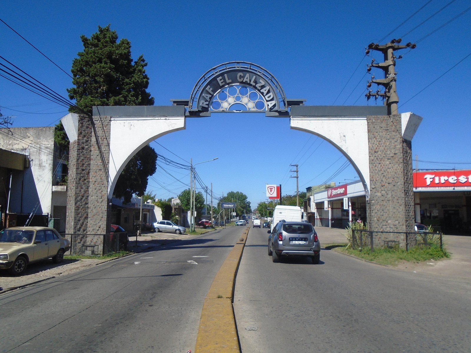 El arco de entrada a Rafael Calzada, con el nombre de la localidad en lo alto, sobre una avenida con autos y comercios a los costados